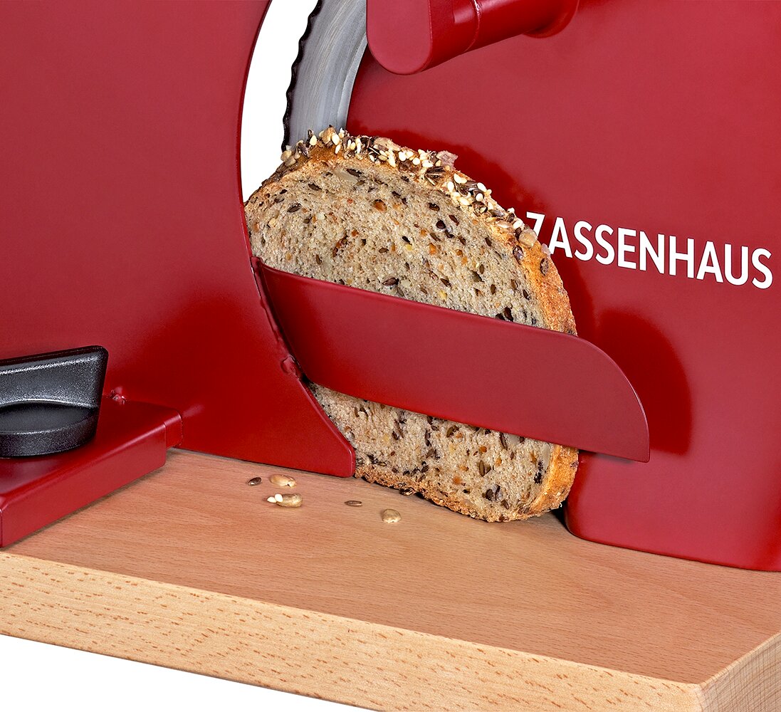 Rote Brotschneidemaschine mit Holzbrett schneidet eine Scheibe Körnerbrot, Nahaufnahme aus seitlicher Perspektive