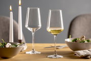 Twee elegante Rieslingglazen op een gedekte tafel, waarvan één gevuld met witte wijn, op de achtergrond brandende kaarsen en schalen met salade, zijaanzicht.
