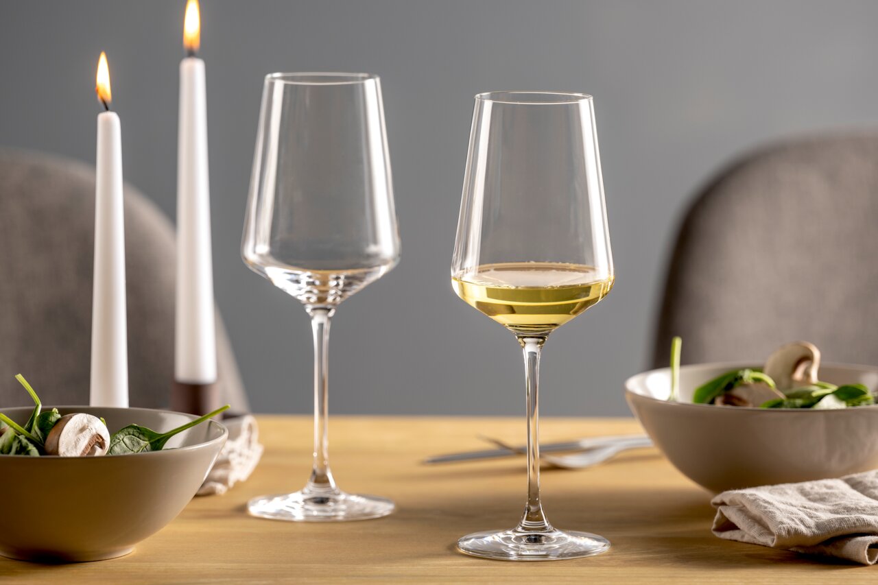 Twee elegante Rieslingglazen op een gedekte tafel, waarvan één gevuld met witte wijn, op de achtergrond brandende kaarsen en schalen met salade, zijaanzicht.