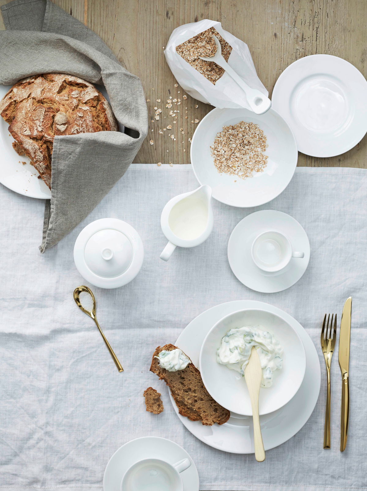 Vogelperspektive auf ein stilvoll gedecktes Frühstückstisch-Arrangement mit weißem Kombiservice, Haferflocken, Brot und goldfarbenem Besteck.