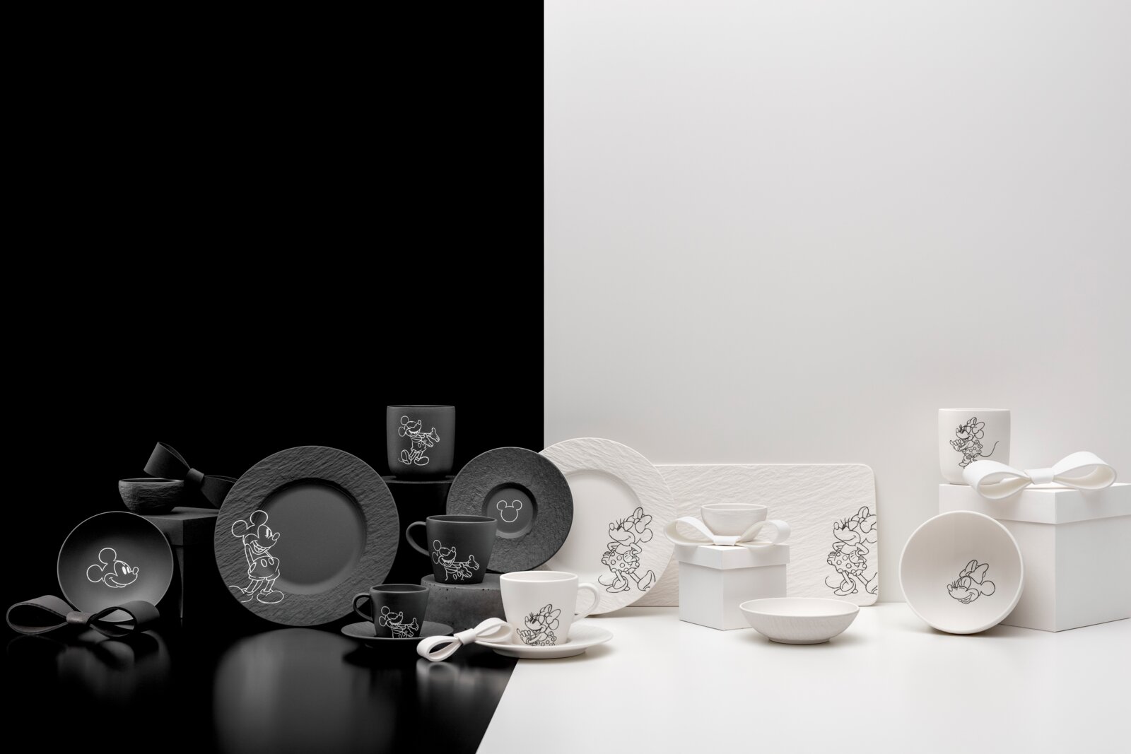 Schwarz-weißes Disney-Tischgeschirr-Set mit Mickey- und Minnie-Motiven (Teller, Tassen, Schalen) neben Geschenkboxen mit Schleifen, frontal in leicht erhöhter Perspektive auf zweifarbigem Studiohintergrund präsentiert.