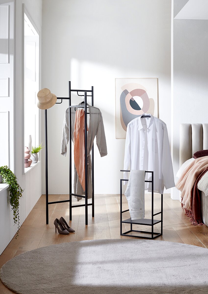 Heller Schlafbereich mit schwarzem Metall-Kleiderständer und passendem Kleiderstuhl, behängt mit Hemd, Kleid, Schal und Hut; Frontansicht auf die Garderobenmöbel im Raum