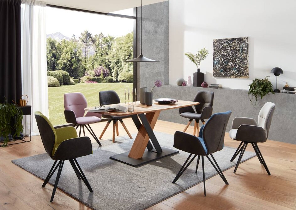 Moderner Essbereich mit einem Esstisch aus Holz und schwarzen Metallbeinen, umgeben von verschiedenen Armlehnstühlen in unterschiedlichen Farben. Die Perspektive zeigt den Raum von der Seite mit Blick auf den Tisch und die Stühle, im Hintergrund ist ein g