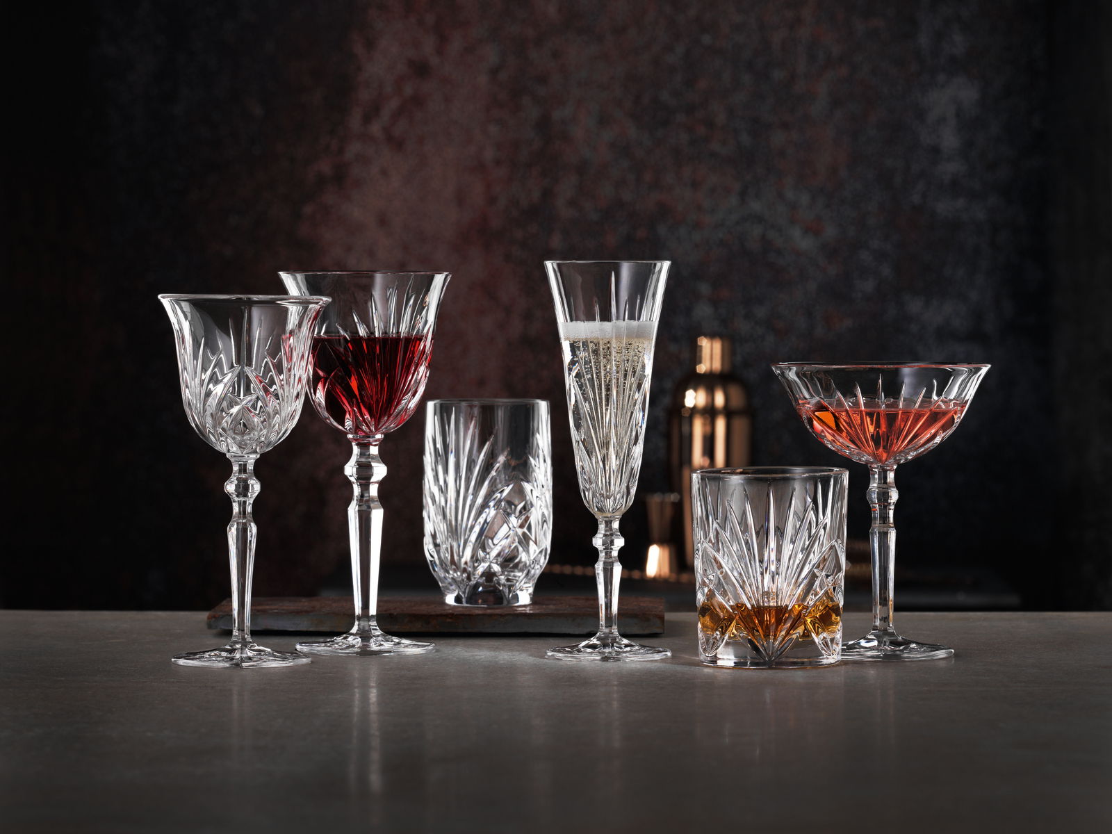 Verschiedene Kristallgläser mit Getränken, frontal auf einem Tisch vor dunklem Hintergrund arrangiert.