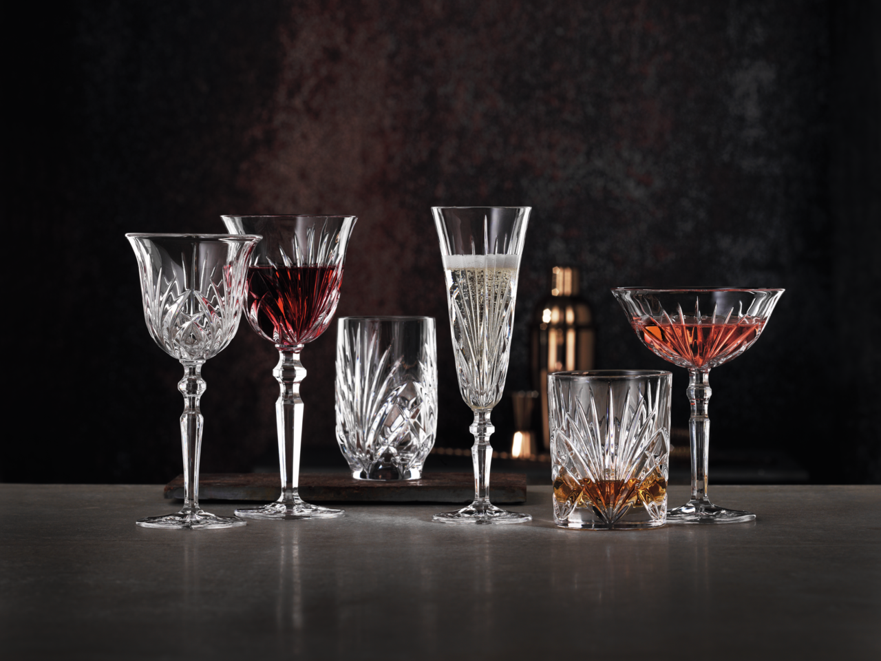 Verschiedene Kristallgläser mit Getränken, frontal auf einem Tisch vor dunklem Hintergrund arrangiert.