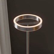 Moderne LED-Stehleuchte mit ringförmigem Leuchtkopf in gebürsteter Metalloptik, warmweiß leuchtend, Detailaufnahme aus leicht seitlicher Perspektive von oben vor dunklem Hintergrund