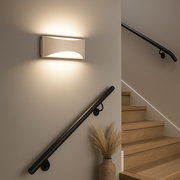 Moderne weiße Wandleuchte im Treppenhaus, die warmes Licht nach oben und unten abstrahlt; Perspektive: seitliche Frontansicht auf die Leuchte an der Wand neben der Treppe.