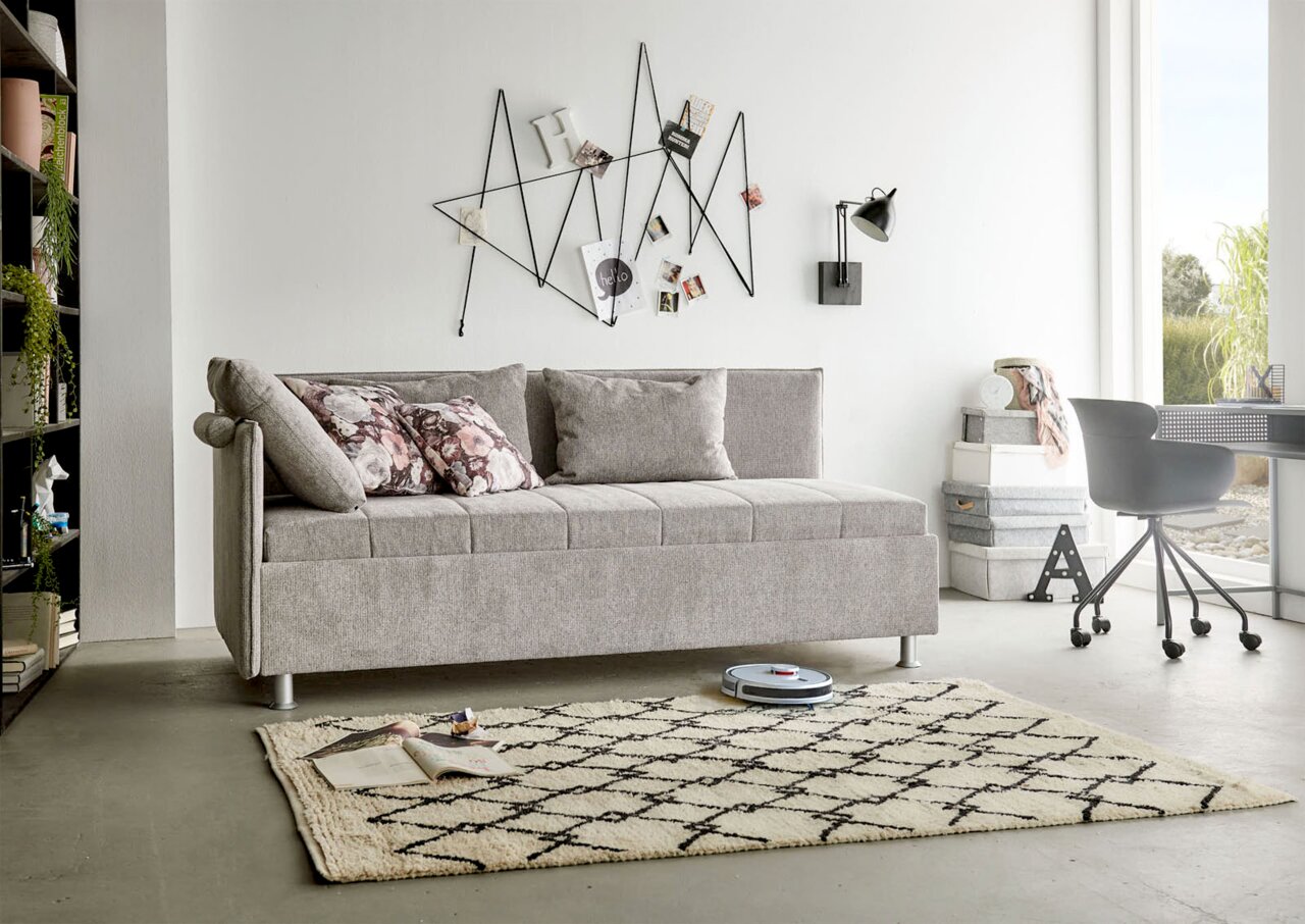 Beldomo Sleep Slaapbank zonder ombouw SANDRO Grijze bank in een moderne woonkamer, gezien vanaf de zijkant, met sierkussens en geometrische wanddecoratie.