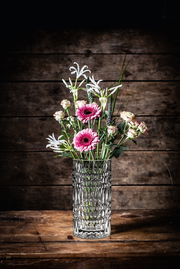 Kristallvase mit Blumenstrauß, frontal auf einem Holztisch vor Holzwand fotografiert.