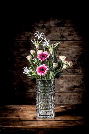Kristallvase mit Blumenstrauß, frontal auf einem Holztisch vor Holzwand fotografiert.