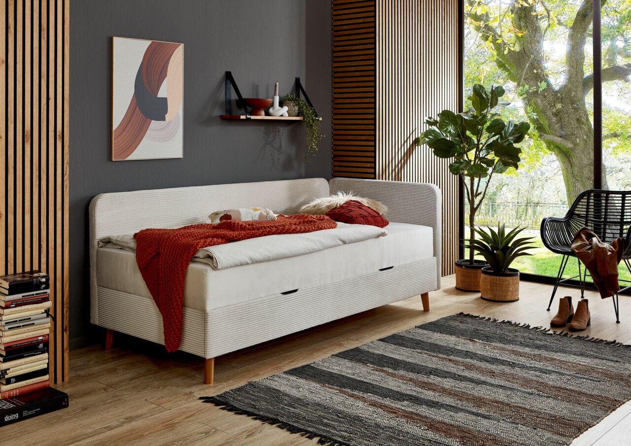 Modernes Polsterbett in einem stilvollen Raum mit Holzboden, dekoriert mit Pflanzen und Kunstwerken, seitliche Perspektive.