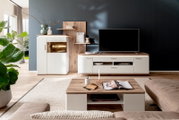 Modernes Wohnzimmer mit weißer Wohnwand und TV-Board in Holzoptik, integriertem Regal und beleuchteter Vitrine neben großem Flachbildfernseher; frontale Perspektive.