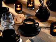 Schwarze Espressotasse mit Untertasse und Latte-Art auf Holztisch, daneben Wasserglas, Gebäck und Kerzenhalter; Perspektive: leicht von oben, Nahaufnahme.