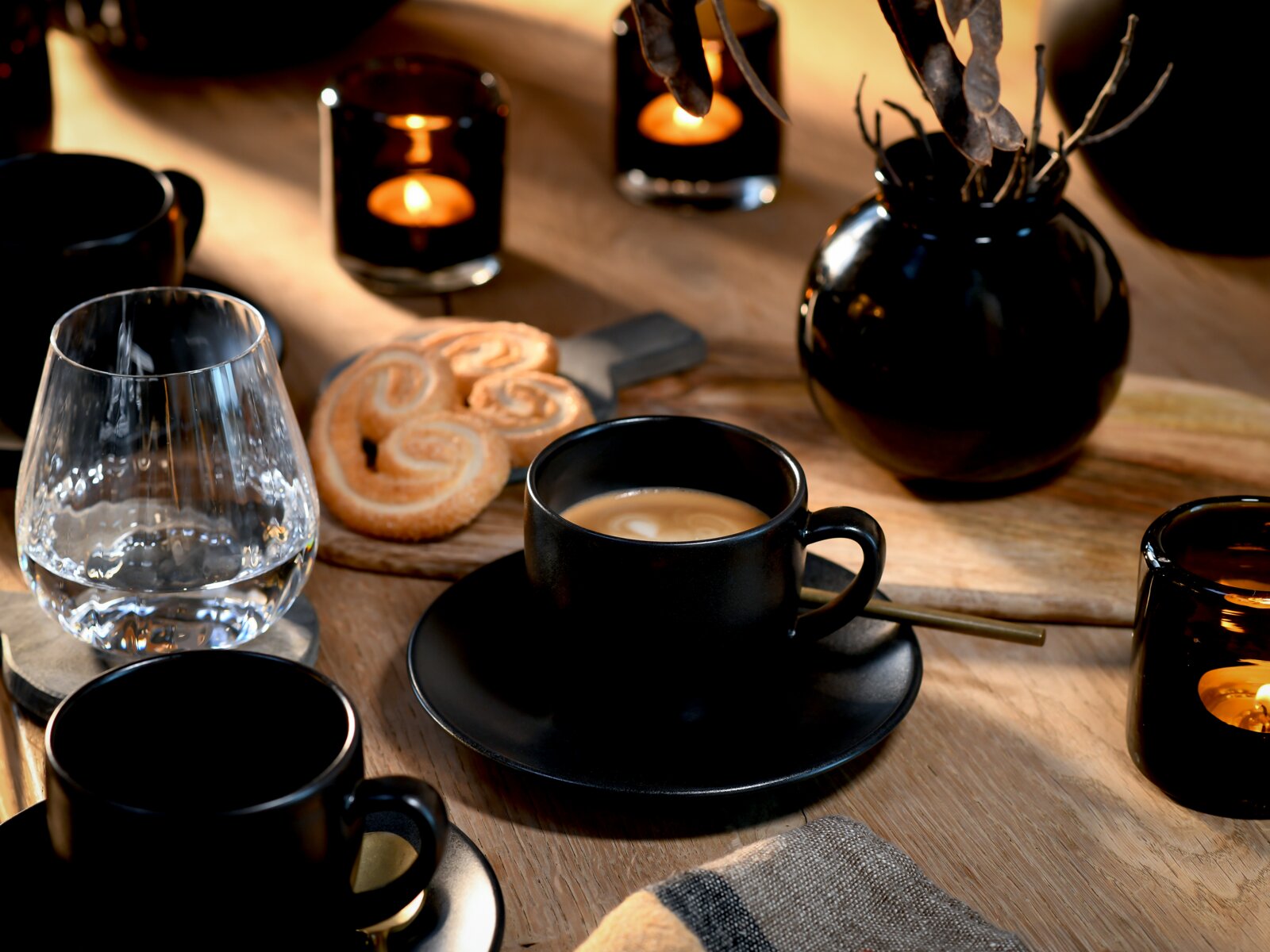 Schwarze Espressotasse mit Untertasse und Latte-Art auf Holztisch, daneben Wasserglas, Gebäck und Kerzenhalter; Perspektive: leicht von oben, Nahaufnahme.