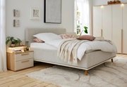 Gemütliches Polsterbett in einem hellen Schlafzimmer, seitliche Perspektive, mit weichen Kissen und Decken, neben einem Nachttisch mit Pflanzen und Büchern.
