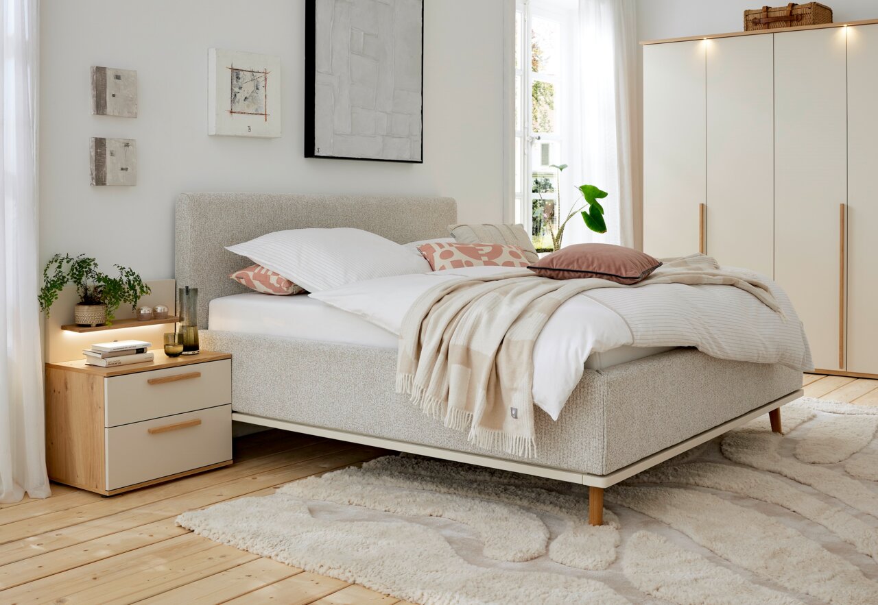 Gemütliches Polsterbett in einem hellen Schlafzimmer, seitliche Perspektive, mit weichen Kissen und Decken, neben einem Nachttisch mit Pflanzen und Büchern.