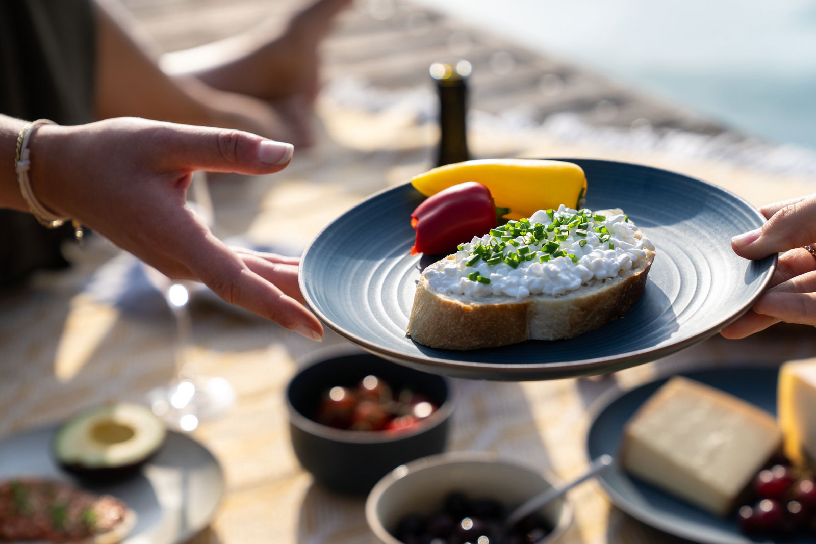 Ein blauer Speiseteller mit einer Scheibe Brot, belegt mit Hüttenkäse und Schnittlauch, sowie Paprikastücken. Der Teller wird von zwei Händen gehalten, im Hintergrund ein Picknick-Setting am Wasser.