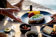 Ein blauer Speiseteller mit einer Scheibe Brot, belegt mit Hüttenkäse und Schnittlauch, sowie Paprikastücken. Der Teller wird von zwei Händen gehalten, im Hintergrund ein Picknick-Setting am Wasser.