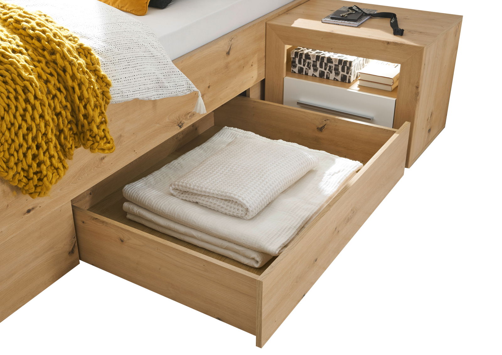 Holzbett mit Schublade, gefüllt mit weißen Decken, und passendem Nachttisch aus Holz, seitliche Perspektive