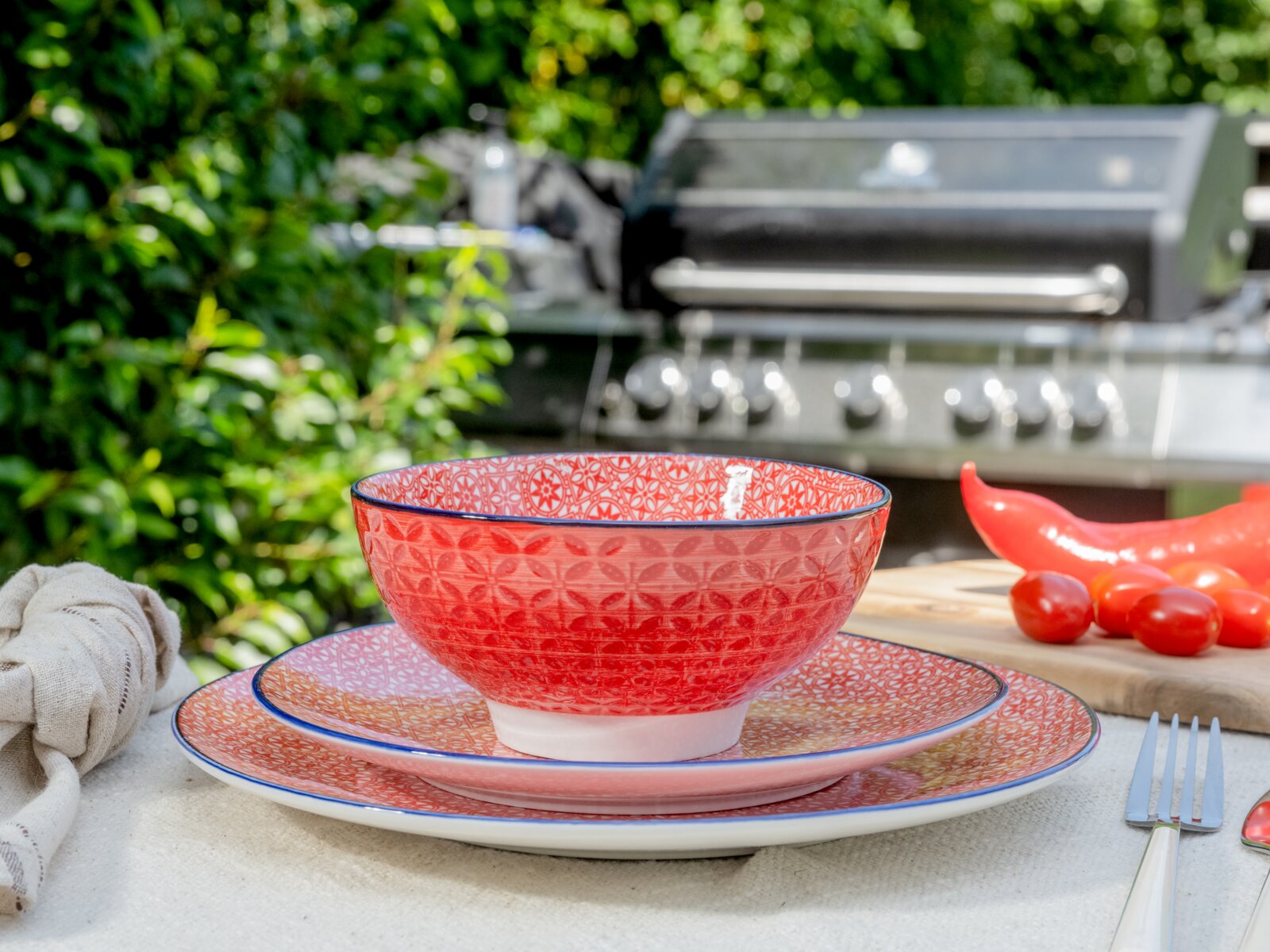 Rot gemusterte Keramikschale auf zwei passenden Tellern auf einem gedeckten Tisch im Garten, seitliche Frontansicht auf Augenhöhe; im Hintergrund Grill, daneben rote Chilis und Tomaten.