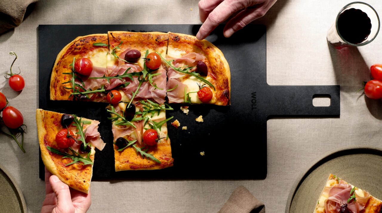 Schneidebrett aus schwarzem Material mit Griff, darauf eine Pizza mit Schinken, Rucola, Tomaten und Oliven, von oben fotografiert.