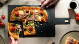 Schneidebrett aus schwarzem Material mit Griff, darauf eine Pizza mit Schinken, Rucola, Tomaten und Oliven, von oben fotografiert.