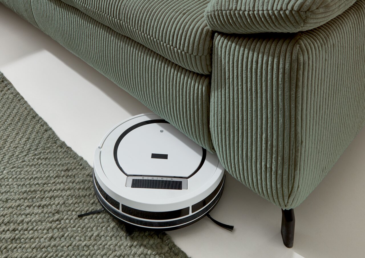 Beldomo Style Ecksofa SENSE Ein Saugroboter reinigt den Boden unter einem grünen Polstersofa aus geripptem Stoff. Perspektive von oben.