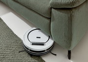 Ein Saugroboter reinigt den Boden unter einem grünen Polstersofa aus geripptem Stoff. Perspektive von oben.