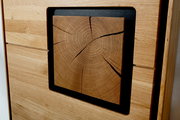Detailaufnahme in leichter Schrägansicht: quadratisches Holz-Element mit sichtbaren Jahresringen und Rissen, eingefasst in einen schwarzen Rahmen in einer hellen Holzfront.
