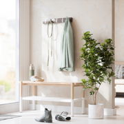 Wandgarderobe mit Haken, an der ein grüner Mantel und eine Leine hängen, daneben eine Bank mit Kopfhörern und einer Trinkflasche, im Vordergrund ein Paar Stiefel und eine grüne Pflanze im Topf, seitliche Perspektive.