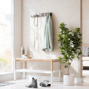 Wandgarderobe mit Haken, an der ein grüner Mantel und eine Leine hängen, daneben eine Bank mit Kopfhörern und einer Trinkflasche, im Vordergrund ein Paar Stiefel und eine grüne Pflanze im Topf, seitliche Perspektive.