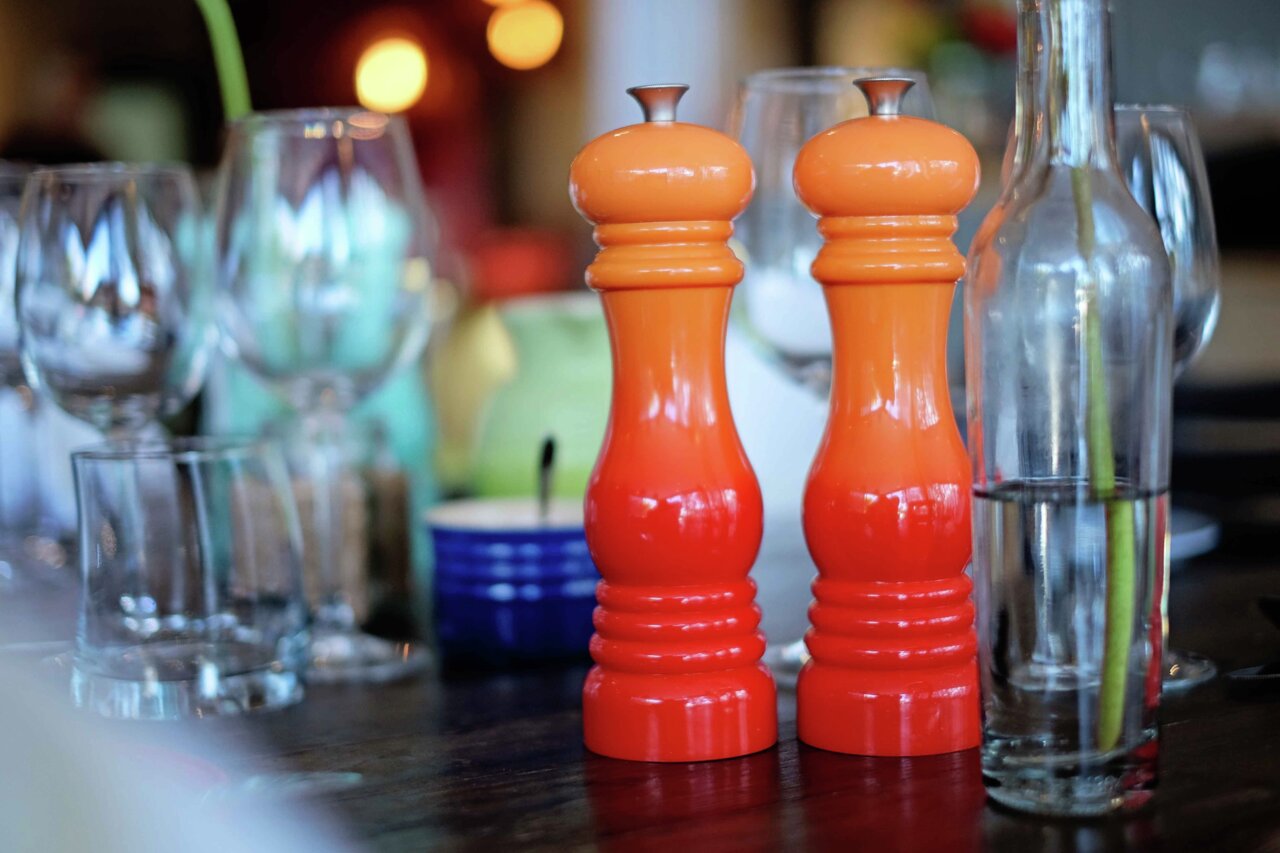Zwei ofenrote Salzmühlen auf einem gedeckten Tisch, umgeben von Gläsern und einer Flasche, aus der Perspektive von vorne.