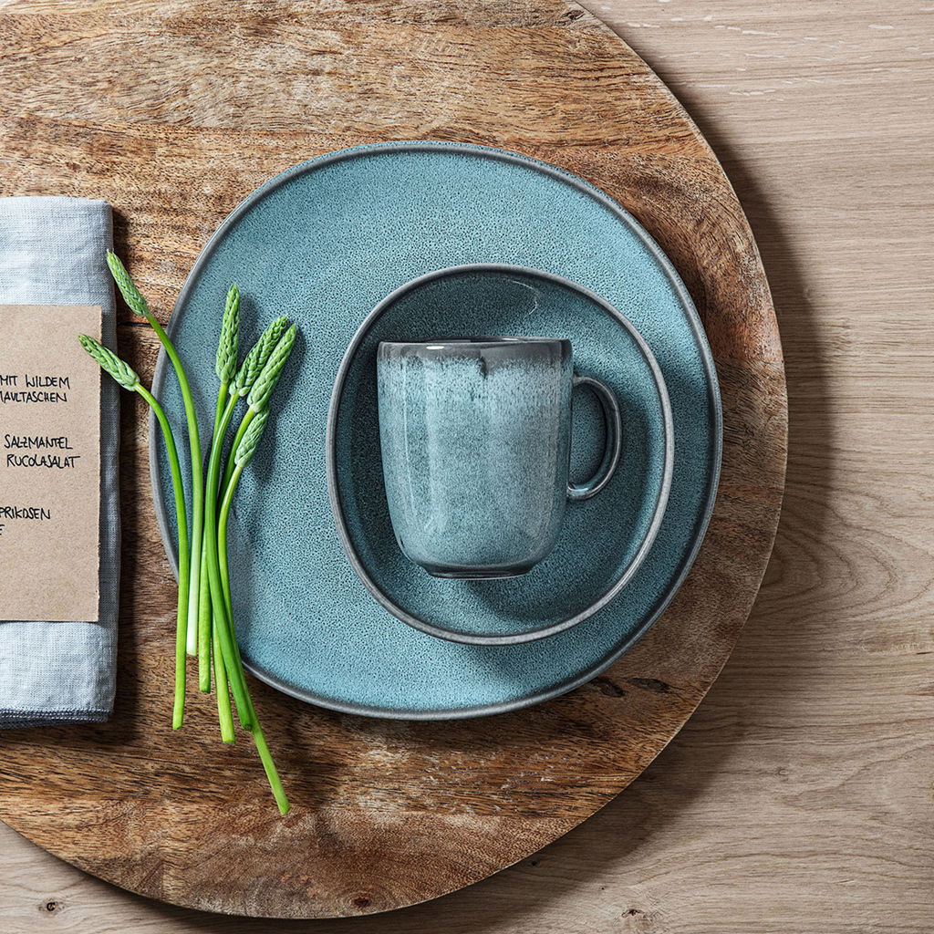 Vogelperspektive auf einen türkisfarbenen Kaffeebecher Lave 340ml, platziert auf einem passenden Teller-Set mit rustikalem Holzuntergrund und frischem grünem Spargel als Dekoration.