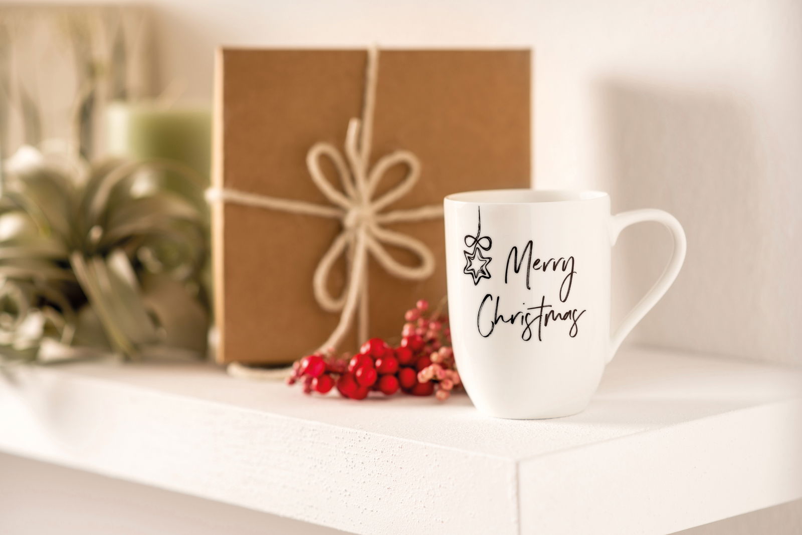 Weiße Tasse mit der Aufschrift 'Merry Christmas' und einem Sternmotiv, auf einem Regal neben einem Geschenk und roten Beeren, seitliche Perspektive.