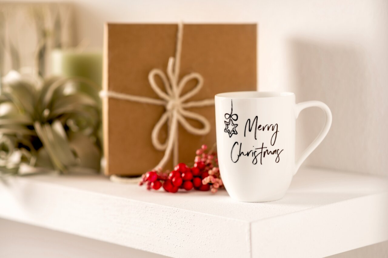 Weiße Tasse mit der Aufschrift 'Merry Christmas' und einem Sternmotiv, auf einem Regal neben einem Geschenk und roten Beeren, seitliche Perspektive.