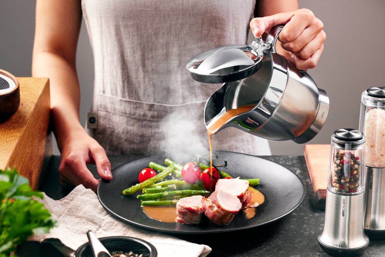 Person gießt Soße aus einer Thermo-Sauciere SERVIRO auf ein Gericht mit Fleisch, grünen Bohnen und Tomaten, seitliche Perspektive.