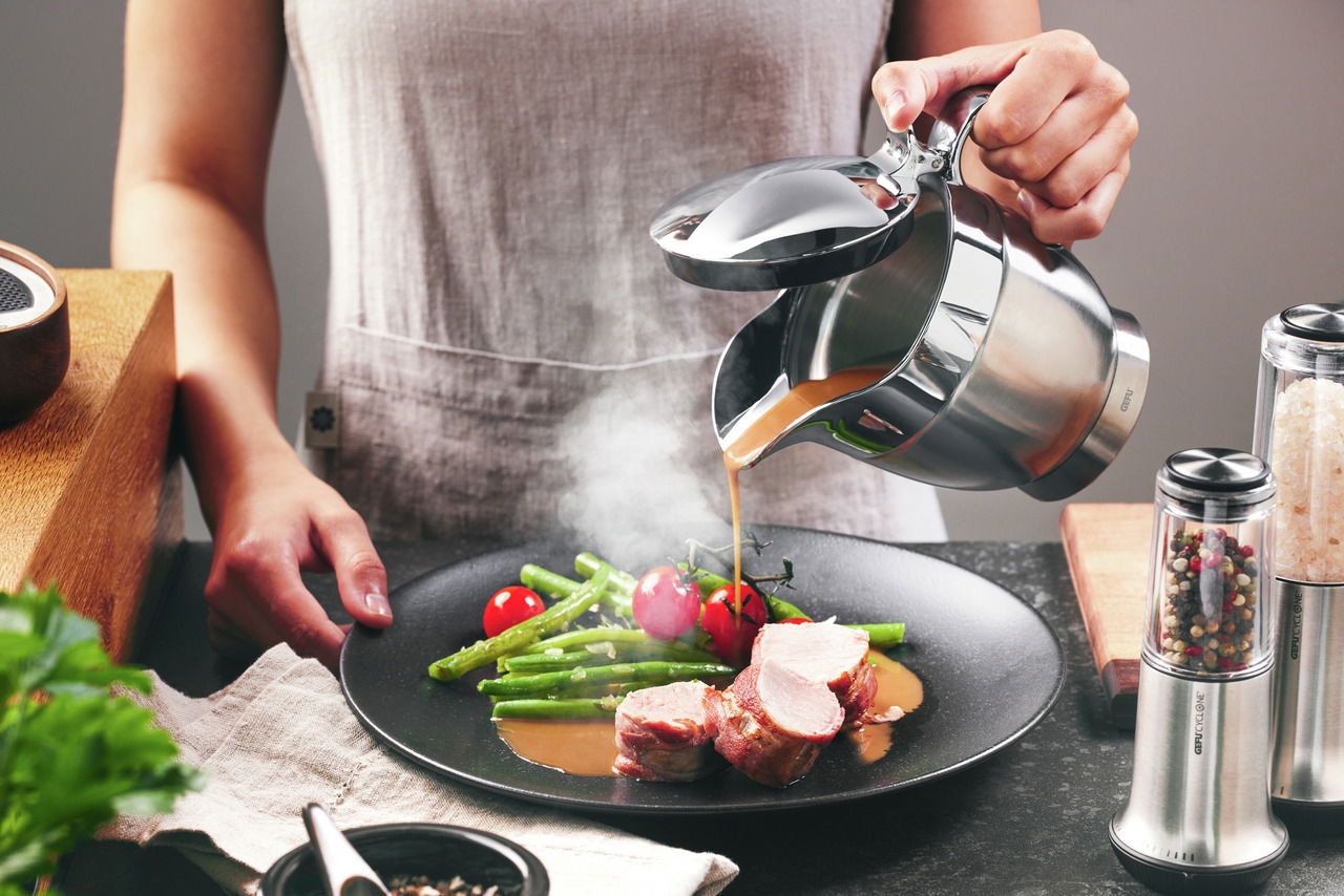 Person gießt Soße aus einer Thermo-Sauciere SERVIRO auf ein Gericht mit Fleisch, grünen Bohnen und Tomaten, seitliche Perspektive.