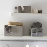 Ein rechteckiger, beige-farbener Korb mit einem Fassungsvermögen von 2 Litern, platziert auf einem Regal. Der Korb hat ein geflochtenes Design und ist von vorne sichtbar. Neben dem Korb liegen gefaltete Kleidungsstücke.