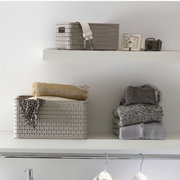 Een rechthoekige, beige mand met een inhoud van 2 liter, die op een plank staat. De mand heeft een gevlochten patroon en is vanaf de voorkant te zien. Naast de mand liggen opgevouwen kledingstukken.
