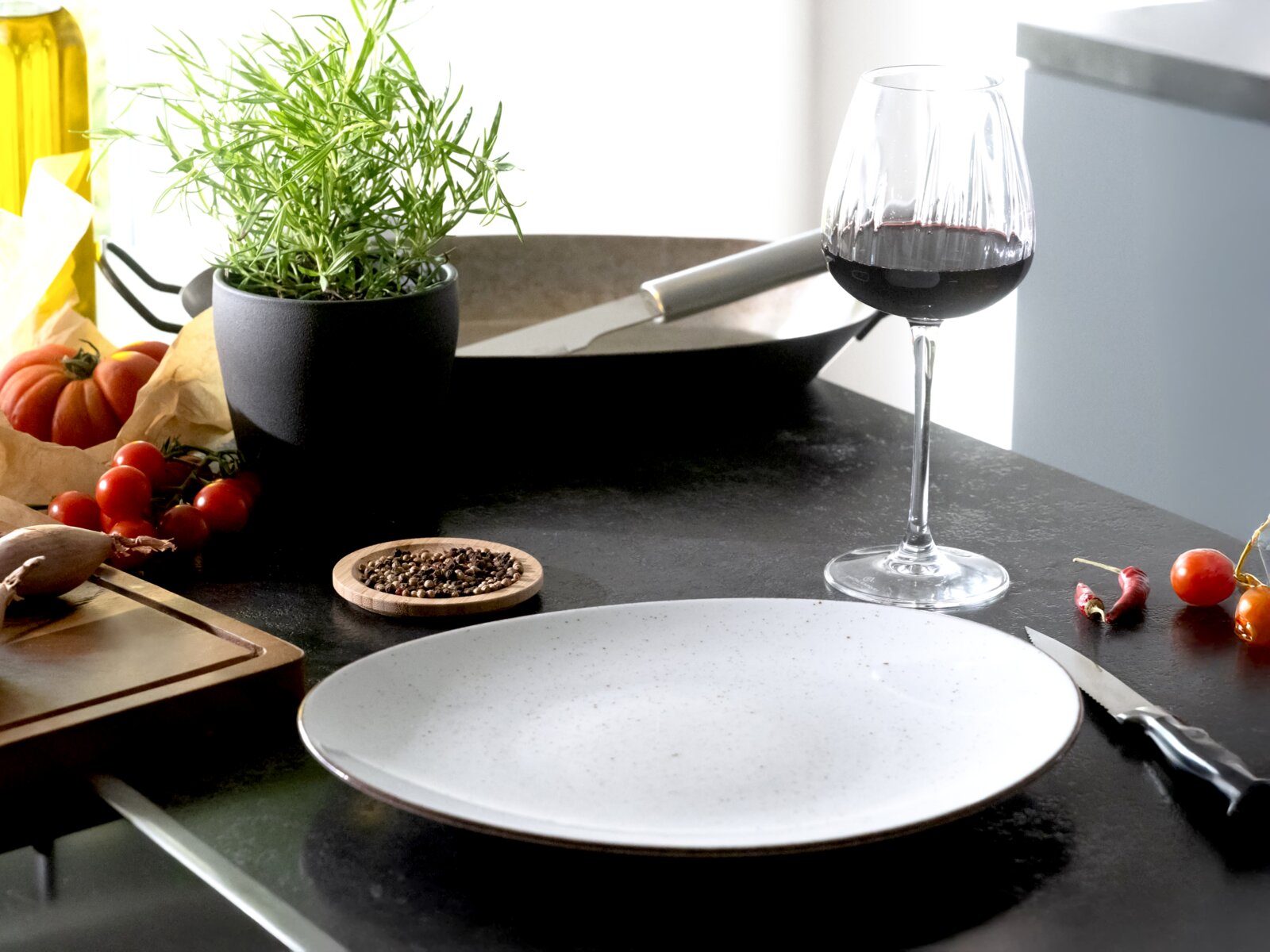 Modern gedeckter Küchentisch mit großem weiß gesprenkeltem Teller im Vordergrund, Weinglas mit Rotwein, schwarzem Topf mit Kräutern und Kochutensilien; Perspektive: leicht seitliche Ansicht auf Tischhöhe.