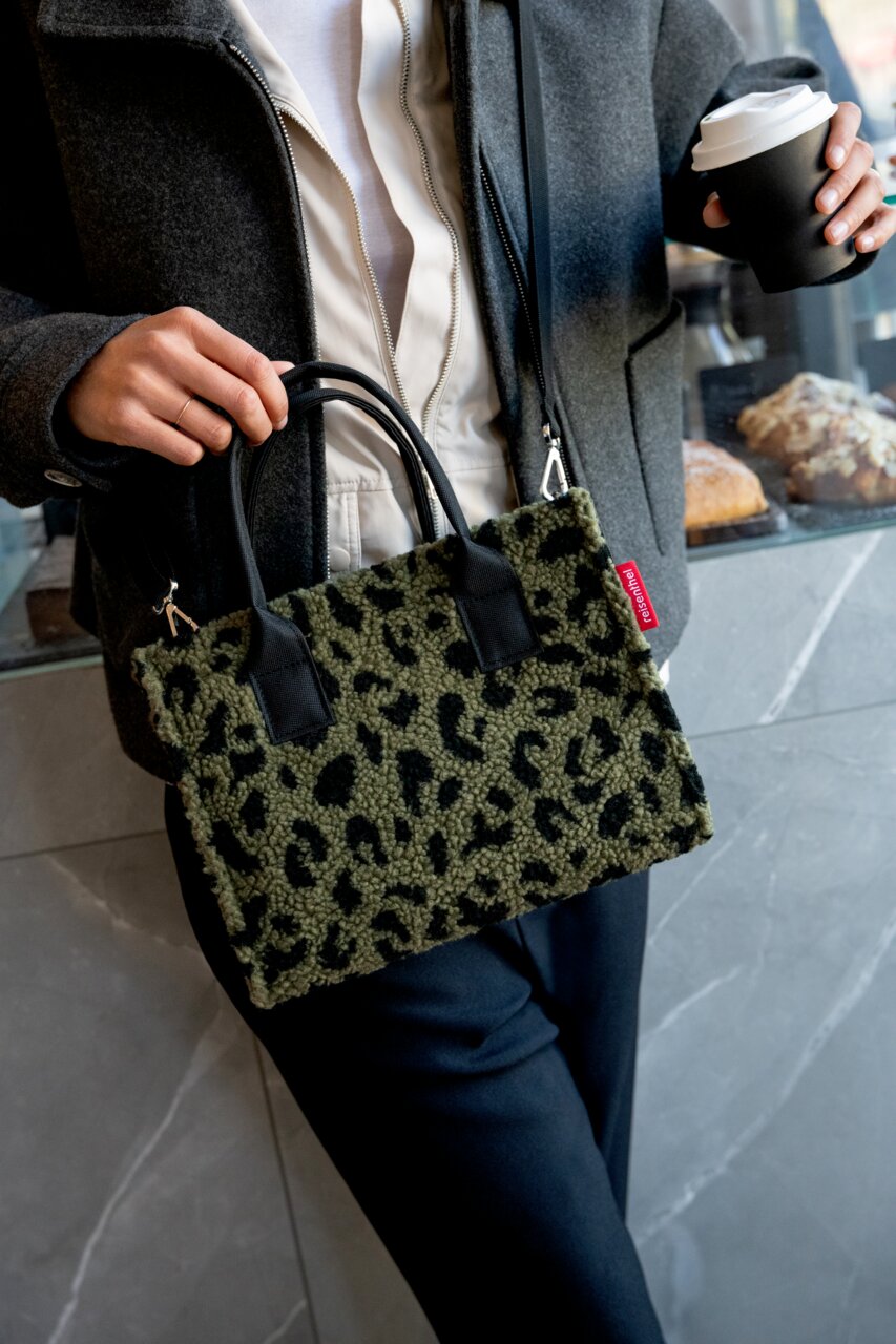Frau hält eine kleine Tasche mit Leopardenmuster in der Hand, während sie einen Kaffee in der anderen Hand hält. Die Tasche hat schwarze Griffe und einen roten Markenanhänger. Perspektive: Frontalansicht.