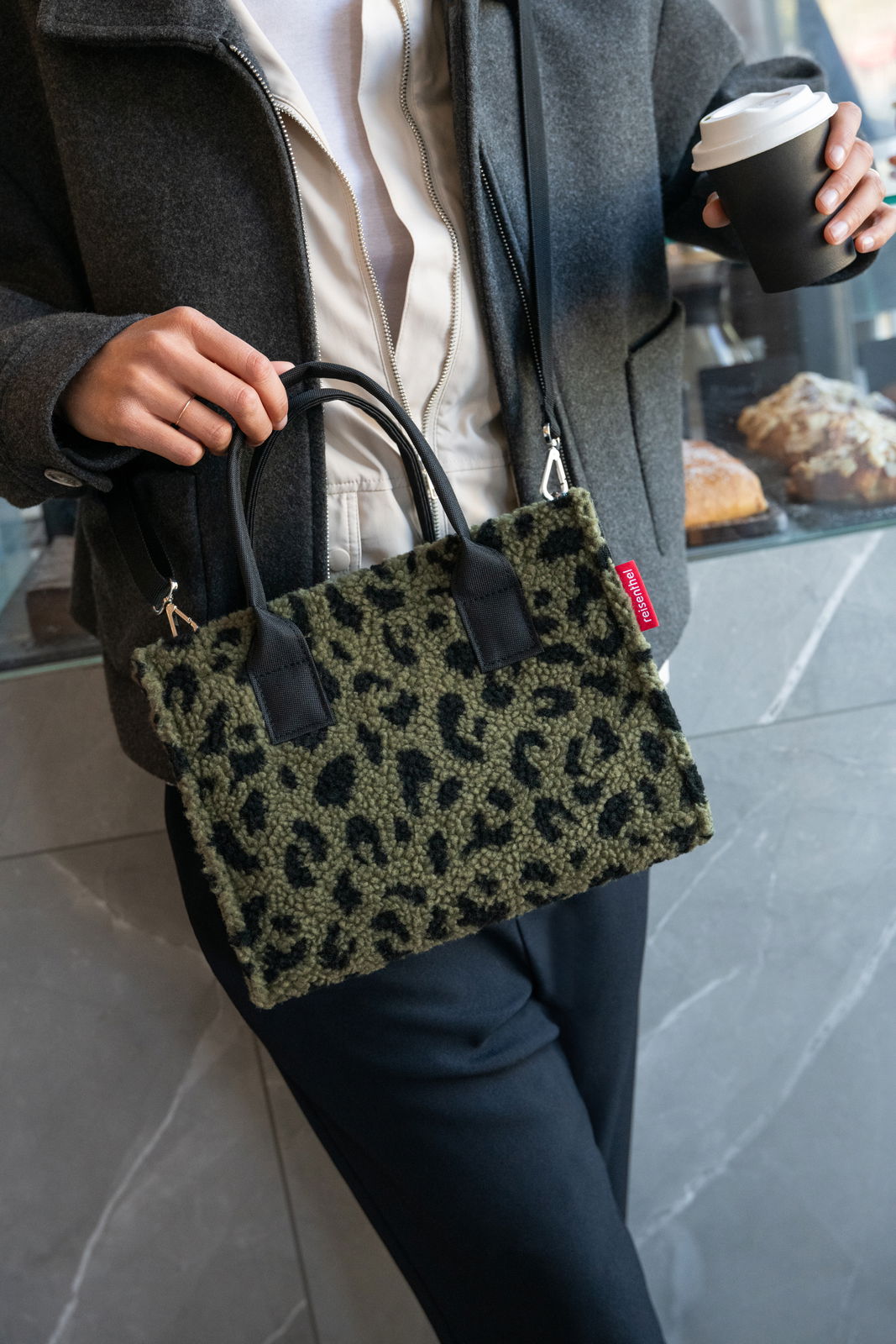Frau hält eine kleine Tasche mit Leopardenmuster in der Hand, während sie einen Kaffee in der anderen Hand hält. Die Tasche hat schwarze Griffe und einen roten Markenanhänger. Perspektive: Frontalansicht.