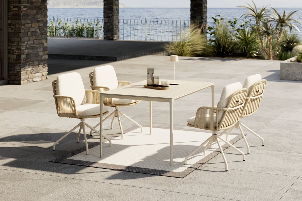 Dieter Knoll Armlehnstuhl DUE Outdoor-Essgruppe mit hellem rechteckigem Tisch und vier gepolsterten, geflochtenen Drehstühlen auf einer modernen Terrasse am Meer, aufgenommen in leichter Schrägansicht von vorne.