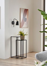 Minimalistischer Beistelltisch aus schwarzem Metall mit quadratischer Tischplatte und offenem Gestell, mit Pflanze dekoriert, in hellem Wohnraum neben Wandlampe und Bild – Perspektive: leicht schräg von vorne auf Augenhöhe.