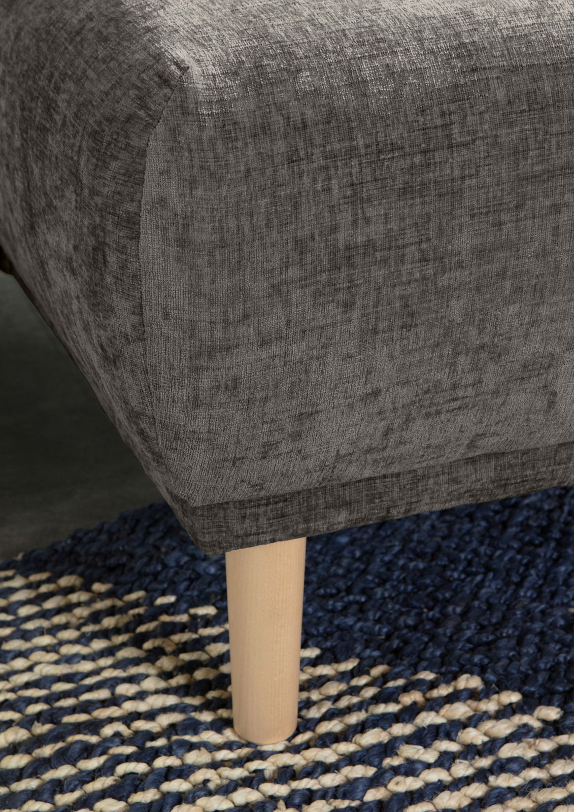 Nahaufnahme einer grauen Rundecke mit Holzbein auf einem blau-weißen Teppich, seitliche Perspektive