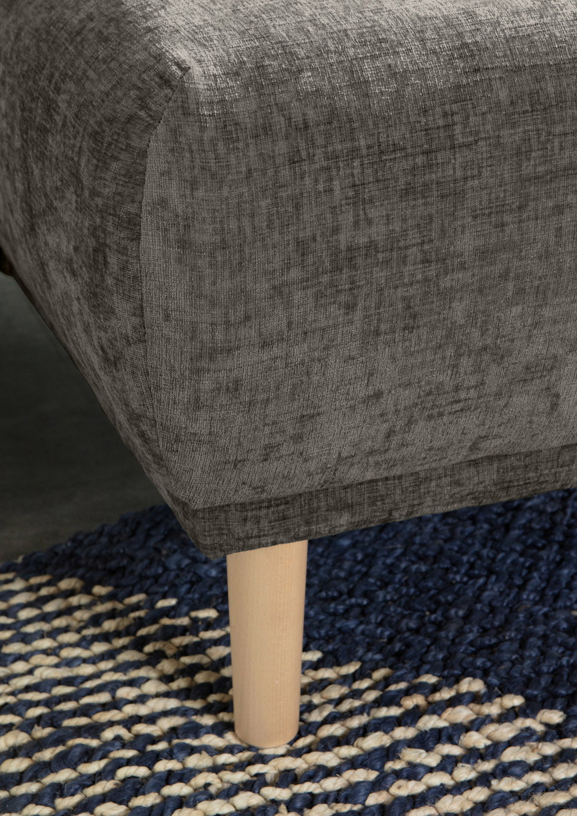 Nahaufnahme einer grauen Rundecke mit Holzbein auf einem blau-weißen Teppich, seitliche Perspektive