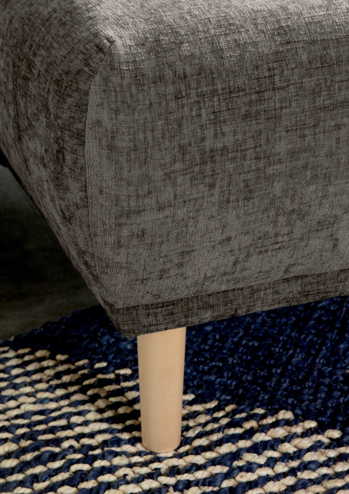 Nahaufnahme einer grauen Rundecke mit Holzbein auf einem blau-weißen Teppich, seitliche Perspektive