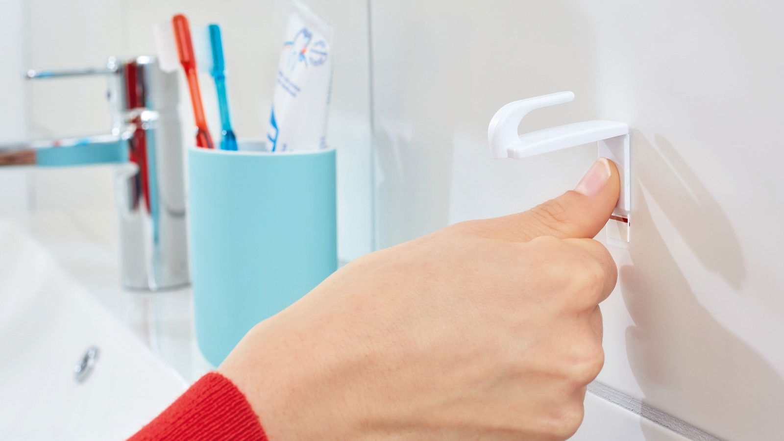 Nahaufnahme einer Hand, die einen weißen tesa Powerstrips Haken Small an einer Badezimmerwand befestigt. Im Hintergrund sind ein Wasserhahn und Zahnbürsten zu sehen.