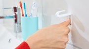 Close-up van een hand die een witte tesa Powerstrips-haak (Small) aan de badkamermuur bevestigt. Op de achtergrond zijn een kraan en tandenborstels te zien.