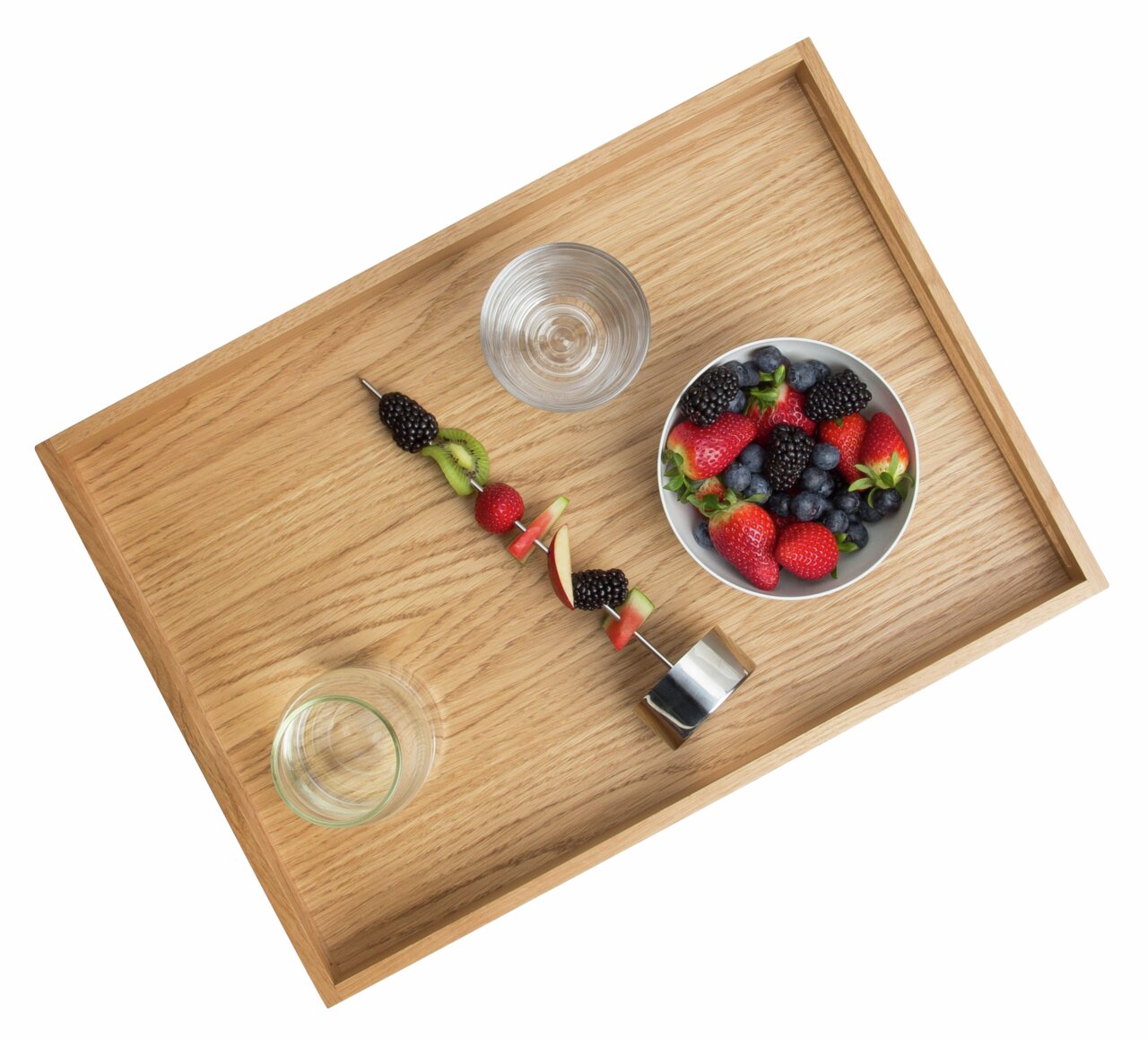 Holztablett aus Eiche mit einer Schale voller Beeren, einem Glas Wasser und einem Fruchtspieß, aus der Vogelperspektive.
